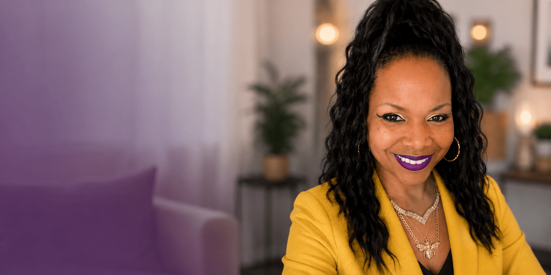 A smiling woman with dark curly hair wearing a bright yellow blazer and purple lipstick.